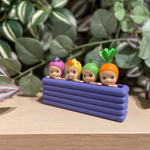 May include: Four small, round-faced figurines with vegetable-themed hats, including a purple hat, a yellow duck hat, a green hat, and an orange carrot hat, are arranged in a purple rectangular planter. The background features green and purple foliage.