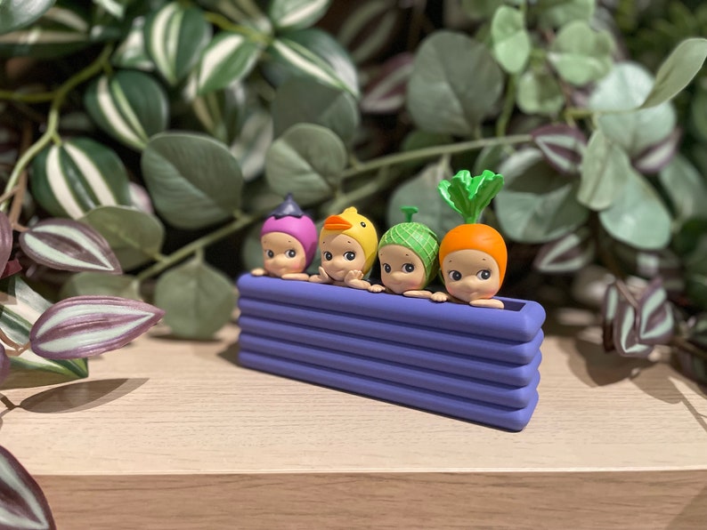 May include: Four small, round-faced figurines with vegetable-themed hats, including a purple hat, a yellow duck hat, a green hat, and an orange carrot hat, are arranged in a purple rectangular planter. The background features green and purple foliage.