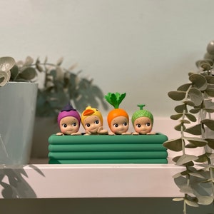 以下が含まれることがあります： 緑色のプランターの縁から、果物や野菜をテーマにした帽子をかぶった4つの小さな気まぐれな置物が見えています。 置物には笑顔があり、一列に並んでいます。 プランターは白い棚の上にあります。