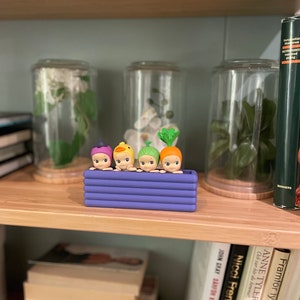 以下が含まれることがあります： 野菜をテーマにした帽子をかぶった、4つの小さな気まぐれなフィギュアが入った紫色の長方形のプランター。フィギュアは笑顔で、一列に並んでいます。プランターは、本やガラス瓶のある木製の棚に置かれています。