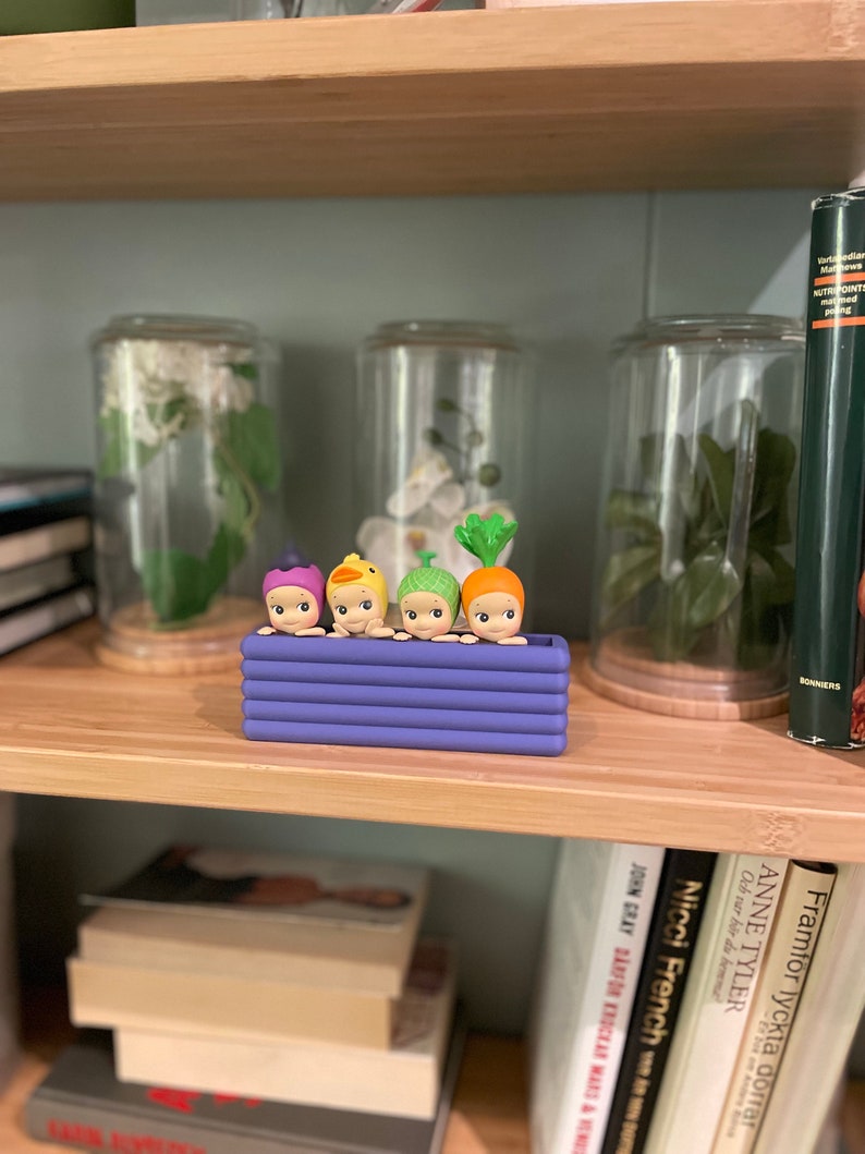 以下が含まれることがあります： 野菜をテーマにした帽子をかぶった、4つの小さな気まぐれなフィギュアが入った紫色の長方形のプランター。フィギュアは笑顔で、一列に並んでいます。プランターは、本やガラス瓶のある木製の棚に置かれています。
