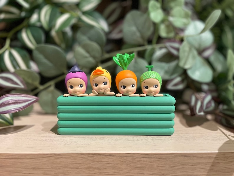 May include: Four small, whimsical figurines with different head coverings, including a witch's hat, a duck, a carrot, and a green-knitted cap, are arranged in a green rectangular planter. The figurines have a light skin tone and are set against a backdrop of green foliage.
