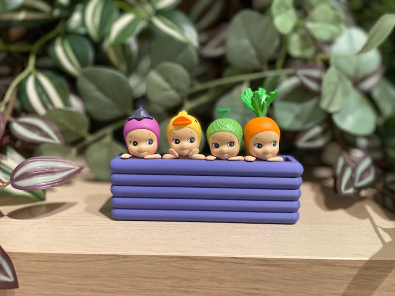 May include: A purple planter box holds four small, whimsical figurines. Each figurine has a different hat: a purple berry, a yellow duck, a green yarn ball, and an orange carrot. The figurines have smiling faces and are a charming decorative item.
