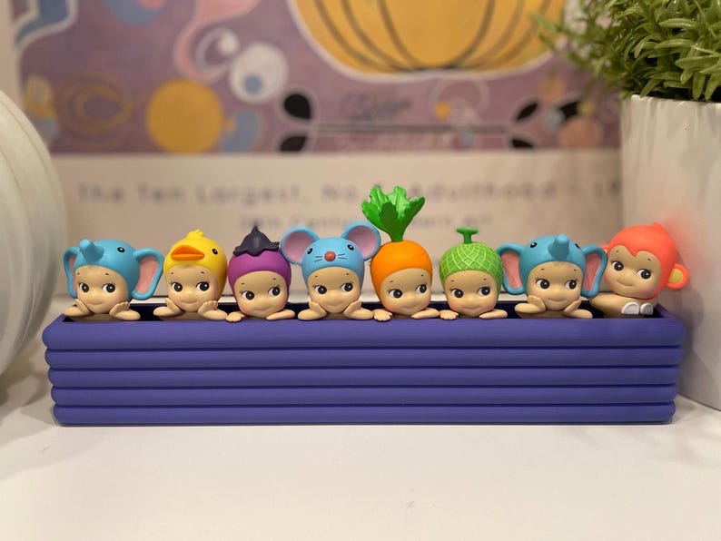 以下が含まれることがあります： 紫色の容器に入った小さなフィギュアのコレクション。象、アヒル、ネズミ、ニンジン、猿など、さまざまな動物や野菜の衣装を着ています。背景にはカラフルな抽象的なデザインがあります。