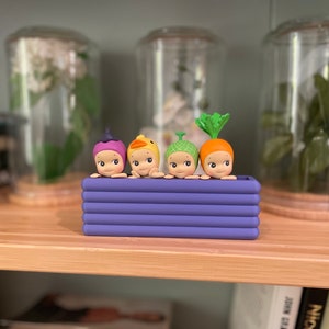 以下が含まれることがあります： 紫色のプランターに入った4体の小さなフィギュアのコレクション。各フィギュアは、紫色のベリー、黄色のアヒル、緑色の芽、オレンジ色のニンジンなど、異なる帽子をかぶっています。フィギュアは、背景にガラス瓶のある木製の棚に置かれています。