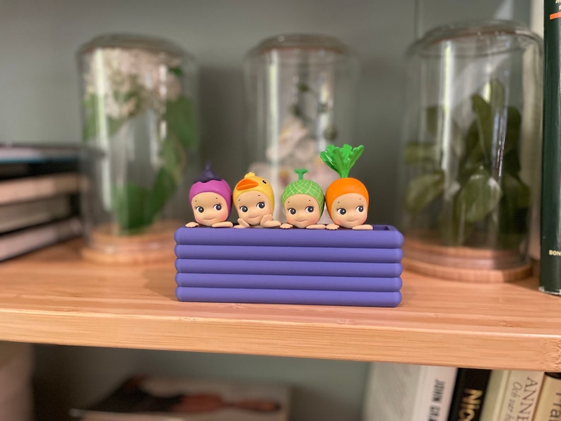 以下が含まれることがあります： 紫色のプランターに入った4体の小さなフィギュアのコレクション。各フィギュアは、紫色のベリー、黄色のアヒル、緑色の芽、オレンジ色のニンジンなど、異なる帽子をかぶっています。フィギュアは、背景にガラス瓶のある木製の棚に置かれています。