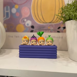 May include: A collection of four small, whimsical figurines in a purple planter. Each figurine has a unique vegetable or animal-themed headpiece, including a duck, onion, carrot, and a green-topped vegetable. The figurines are set against a blurred background.