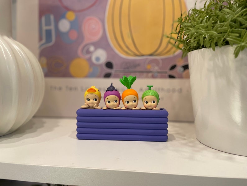 May include: A collection of four small, whimsical figurines in a purple planter. Each figurine has a unique vegetable or animal-themed headpiece, including a duck, onion, carrot, and a green-topped vegetable. The figurines are set against a blurred background.