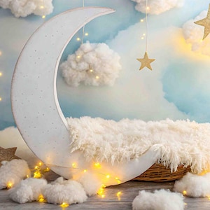 May include: A white crescent moon shaped backdrop with a fluffy white rug inside. The backdrop is decorated with white clouds, gold glitter stars, and white fairy lights.
