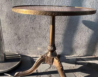 Puede incluir: Una mesa auxiliar redonda de color dorado con un borde decorativo ornamentado en la parte superior. La mesa tiene tres patas con tallas intrincadas y un pedestal central.