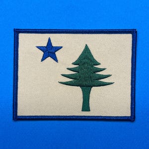 May include: Embroidered patch featuring the Maine state flag design. The patch has a cream-colored background with a blue star and a green pine tree. A blue border frames the design.