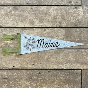 Puede incluir: Un banderín de fieltro gris con la palabra "Maine" en escritura cursiva. Presenta una ilustración negra de un pájaro en una rama de pino con piñas. El banderín tiene detalles de fieltro verde y se muestra sobre una superficie de madera.