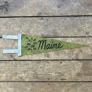 Puede incluir: Bandera de fieltro verde con la palabra "Maine" en escritura negra. Una ilustración negra de un pájaro y piñas decora la bandera. La bandera tiene detalles de fieltro gris y se muestra sobre una superficie de madera.
