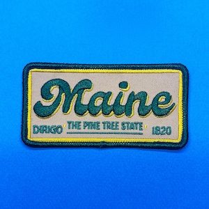 May include: Embroidered patch featuring the word "Maine" in a stylized green font. The patch includes the phrases "DIRIGO" and "THE PINE TREE STATE 1820" in yellow lettering. The patch has a tan background with a yellow border and a dark green stitched edge.
