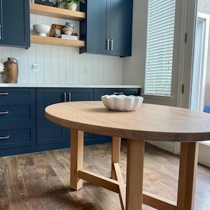 May include: A light wood oval table with a white ceramic bowl on top. The table has a simple, modern design with four legs and a cross-shaped support. The table is in a kitchen with dark blue cabinets and a wood floor.