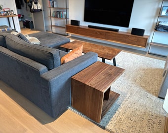 May include: A dark gray sectional sofa with a brown wooden coffee table and a smaller brown wooden side table. The sofa is in a living room with a rug on the floor.