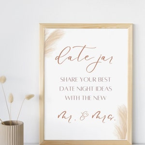 Op de afbeelding: Een wit bord met een houten frame en een bruine inscriptie die "date jar" luidt. Het bord bevat ook de tekst "Share your best date night ideas with the new Mr. & Mrs."