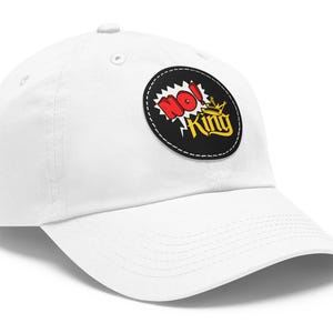 May include: White baseball cap with a curved peak. The cap has a circular patch with the words "NO! King" in red, yellow, and black. The patch is outlined with a black border and a white stitched edge. The cap is made of a soft material.