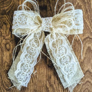 May include: Cream-colored burlap bow with delicate lace detailing. The bow features a layered design with long, fringed tails and a textured, rustic appearance. The bow is set against a warm, wooden background, highlighting its natural tones and handcrafted aesthetic.
