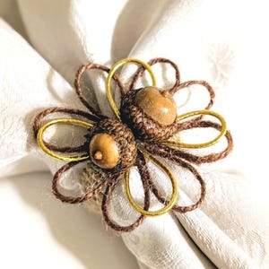 May include: A decorative napkin ring featuring two acorns with brown caps and textured bodies. The acorns are surrounded by a flower-shaped design made of brown twine and gold-colored wire. The napkin ring is resting on a white cloth napkin.