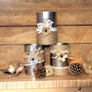 May include: Three upcycled tin cans decorated with twine, white ribbon, and floral accents. The cans are arranged on a wooden surface, with additional decorative elements including a pinecone and a woven ball.