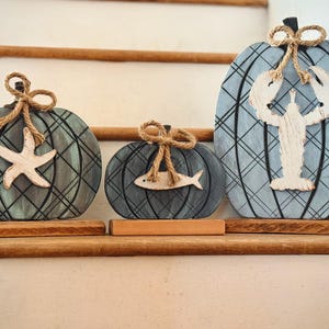 May include: Three flat, pumpkin-shaped wooden decorations with a blue and black plaid pattern. Each pumpkin sits on a rectangular wooden base and features a jute twine bow at the top. The pumpkins are adorned with white wooden cutouts of a starfish, a fish, and a lobster.