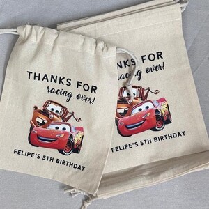 May include: Two beige drawstring bags featuring Lightning McQueen and Mater from the Cars film. The bags have the text "THANKS FOR racing over!" and "FELIPE'S 5TH BIRTHDAY" printed on them.