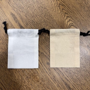 May include: Two small, square drawstring pouches. One is white, the other is beige. Both have a black drawstring closure at the top. The pouches are made of a textured fabric and are displayed on a wooden surface.