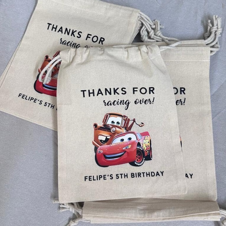 May include: Beige drawstring party bags featuring Lightning McQueen and Mater from the Cars film. The bags have the text "THANKS FOR racing over!" and "FELIPE'S 5TH BIRTHDAY" printed on them.