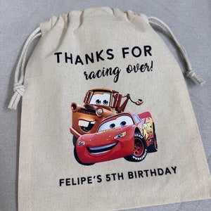 May include: A beige drawstring bag with Lightning McQueen and Mater from Cars. The bag has the text "THANKS FOR racing over!" and "FELIPE'S 5TH BIRTHDAY" printed in black. The bag is made of a canvas-like material.