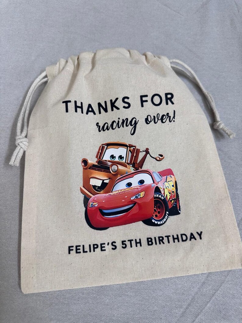 May include: A beige drawstring bag with Lightning McQueen and Mater from Cars. The bag has the text "THANKS FOR racing over!" and "FELIPE'S 5TH BIRTHDAY" printed in black. The bag is made of a canvas-like material.