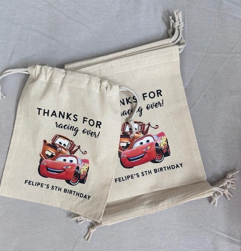 May include: Two beige drawstring bags featuring Lightning McQueen and Mater from the Cars film. The bags have the text "THANKS FOR racing over!" and "FELIPE'S 5TH BIRTHDAY" printed on them.