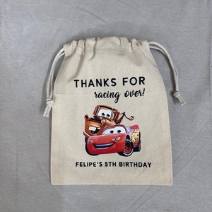 May include: A beige drawstring bag with the text "THANKS FOR racing over!" and "FELIPE'S 5TH BIRTHDAY". The bag displays a colourful graphic of Lightning McQueen and Mater from the Cars movie.