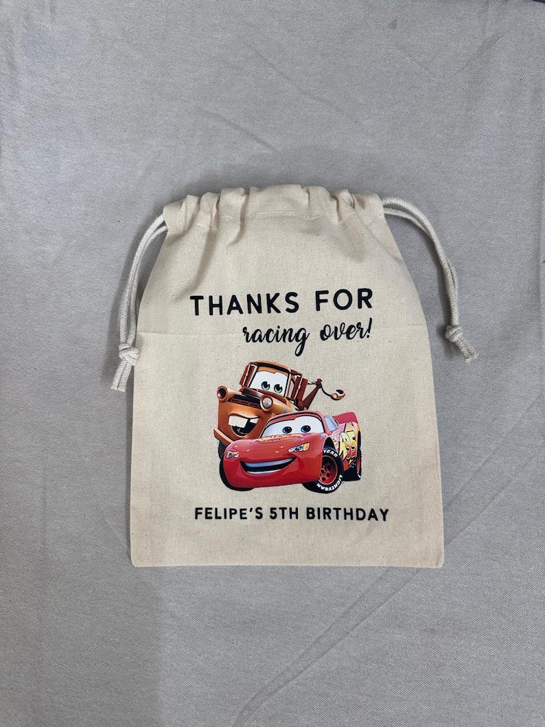 May include: A beige drawstring bag with the text "THANKS FOR racing over!" and "FELIPE'S 5TH BIRTHDAY". The bag displays a colourful graphic of Lightning McQueen and Mater from the Cars movie.