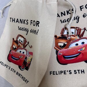 May include: Two beige drawstring bags with a Cars theme. Each bag features Lightning McQueen and Mater, with the text "THANKS FOR racing over!" and "FELIPE'S 5TH BIRTHDAY" printed in black. The bags have a white drawstring closure.