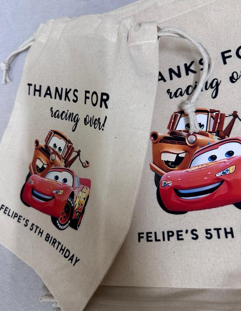 May include: Two beige drawstring bags with a Cars theme. Each bag features Lightning McQueen and Mater, with the text "THANKS FOR racing over!" and "FELIPE'S 5TH BIRTHDAY" printed in black. The bags have a white drawstring closure.