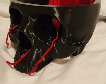 May include: A black skull-shaped yarn bowl with red yarn inside. The yarn is spilling out of the bowl's eye sockets.