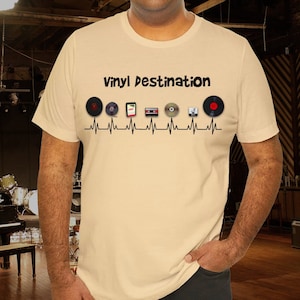 May include: Beige t-shirt with the text "Vinyl Destination" above a series of music media icons, including vinyl records, a cassette tape, and a CD, all connected by a black heartbeat line.