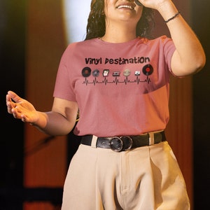 May include: A dusty rose t-shirt with the text "Vinyl Destination" and a graphic of vinyl records, a cassette tape, and a CD. The shirt is worn with tan pants and a black belt with a silver buckle.