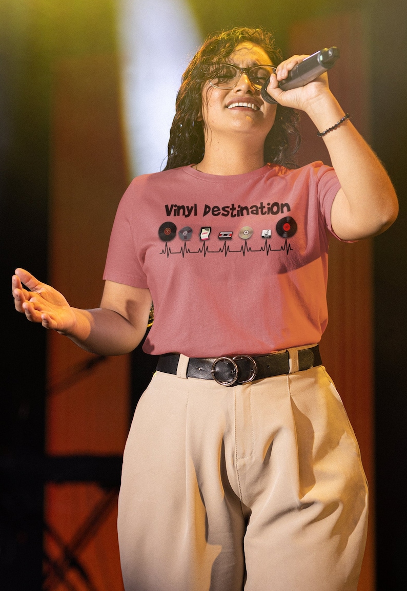 May include: A dusty rose t-shirt with the text "Vinyl Destination" and a graphic of vinyl records, a cassette tape, and a CD. The shirt is worn with tan pants and a black belt with a silver buckle.