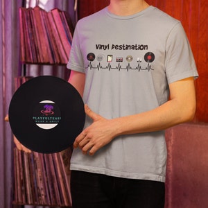 May include: A light blue t-shirt with the text "Vinyl Destination" and a graphic of vinyl records, a cassette tape, and other music-related icons. A black vinyl record is held in front of the shirt. The record has the text "PLAYFULTEASI WEAR A SMILE".