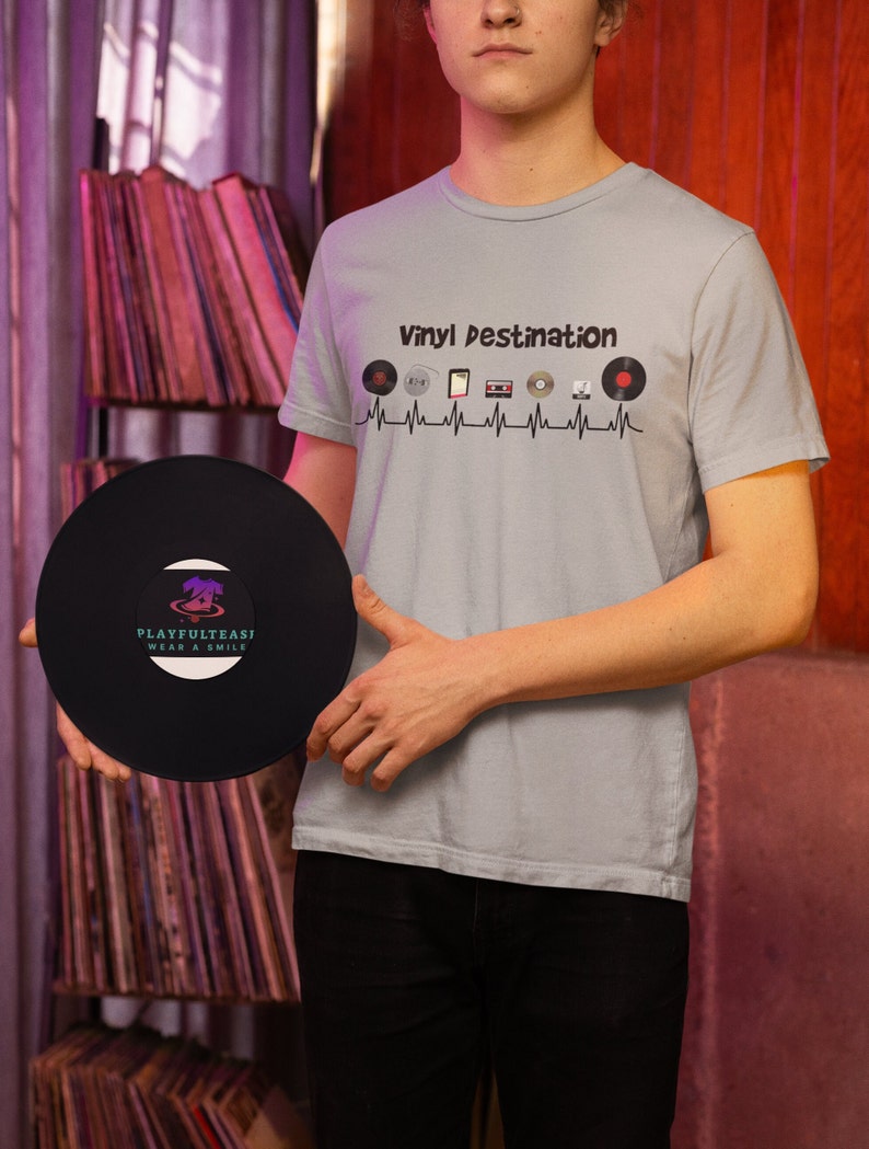 May include: A light blue t-shirt with the text "Vinyl Destination" and a graphic of vinyl records, a cassette tape, and other music-related icons. A black vinyl record is held in front of the shirt. The record has the text "PLAYFULTEASI WEAR A SMILE".