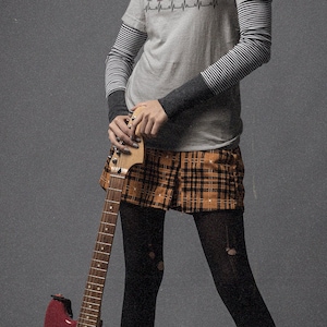May include: A person wearing a gray t-shirt with "Vinyl Destination" text and a heartbeat graphic, layered over a striped long-sleeve shirt, paired with a plaid skirt, black tights, and boots, holding a red electric guitar.