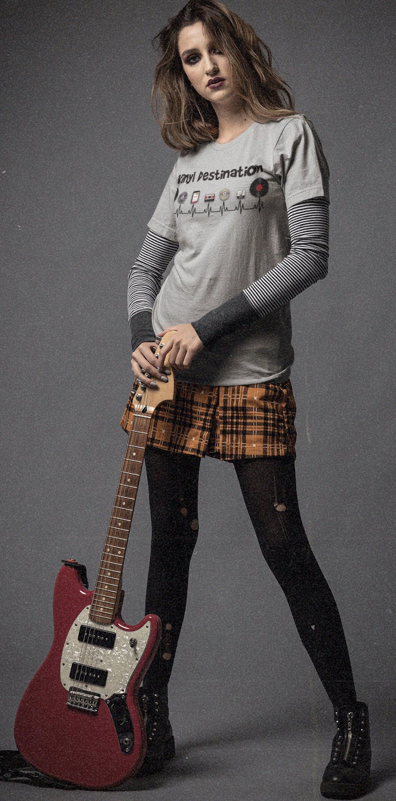 May include: A person wearing a gray t-shirt with "Vinyl Destination" text and a heartbeat graphic, layered over a striped long-sleeve shirt, paired with a plaid skirt, black tights, and boots, holding a red electric guitar.