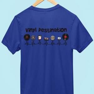 May include: Royal blue t-shirt with the text "VINYL DESTINATION" above a series of vinyl records, cassettes, and CDs. The design is set against a black heartbeat line. The shirt is hanging on a wooden hanger.