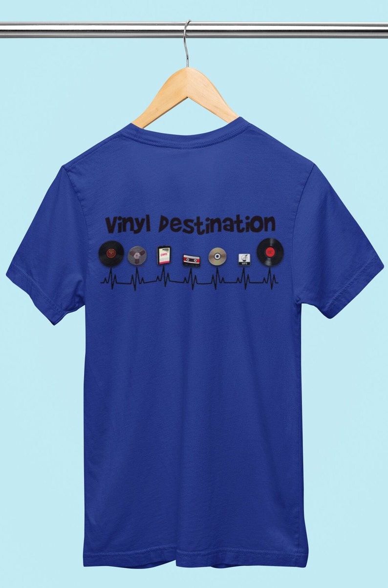 May include: Royal blue t-shirt with the text "VINYL DESTINATION" above a series of vinyl records, cassettes, and CDs. The design is set against a black heartbeat line. The shirt is hanging on a wooden hanger.