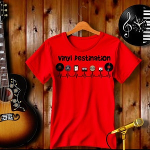 May include: A red t-shirt with the text "Vinyl Destination" and various music-related icons. The shirt is hanging on a wooden hanger. A guitar and a microphone are also in the image.