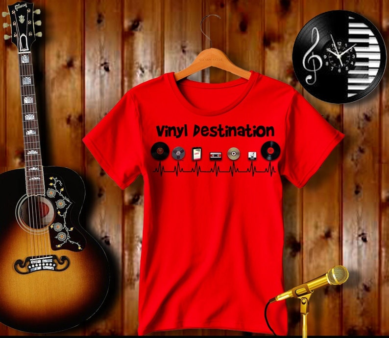 May include: A red t-shirt with the text "Vinyl Destination" and various music-related icons. The shirt is hanging on a wooden hanger. A guitar and a microphone are also in the image.