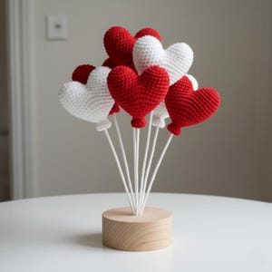 Puede incluir: Una pieza decorativa artesanal con globos en forma de corazón de ganchillo, rojos y blancos. Los globos están sujetos a palitos blancos y colocados en una base de madera. Ideal para San Valentín u otras ocasiones especiales.
