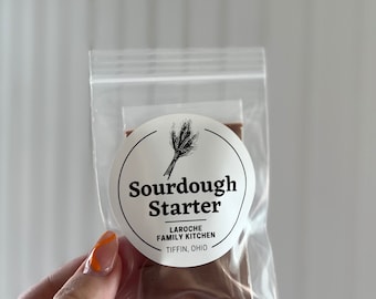 Könnte beinhalten: Ein klarer Plastikbeutel mit einem Sauerteigstarter. Der Beutel hat ein weißes rundes Etikett mit den Worten "Sourdough Starter" und "LAROCHE FAMILY KITCHEN TIFFIN, OHIO" aufgedruckt. Das Etikett zeigt auch eine Weizengrafik.